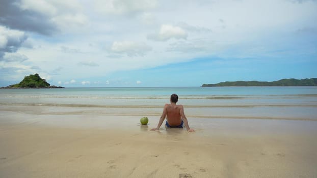 Man sits on a sandy beach and enjoy the seascape. Man relaxing sitting down on the shore of a white sand beach, with the waves bathing the shore in an idyllic nature location, being thoughtful.