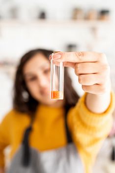 Beautiful brunette woman in yellow sweater and gray apron coloring easter eggs in the kitchen holding test tube with orange color