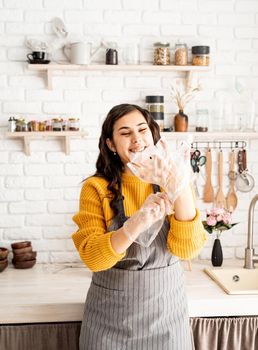 Beautiful brunette laughing woman in yellow sweater and gray apron preparing to color easter eggs in the kitchen putting on the gloves