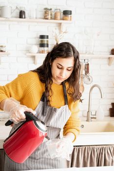 Beautiful brunette woman in yellow sweater and gray apron preparing to color easter eggs in the kitchen, pouring water into the cup