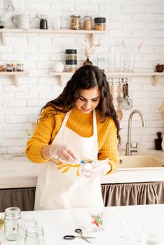 Beautiful brunette woman in yellow sweater and gray apron coloring easter eggs blue in the kitchen