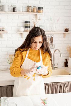 Beautiful brunette woman in yellow sweater and gray apron coloring easter eggs blue in the kitchen