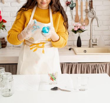 Beautiful brunette woman in yellow sweater and gray apron coloring easter eggs blue in the kitchen
