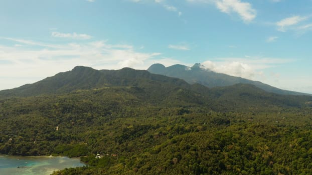 Tropical landscape: Mountains with forest, against blue sea with blue sky with clouds, top view, Camiguin, Philippines. Mountain landscape on tropical island with mountain peaks covered with forest. Slopes of mountains with evergreen vegetation.