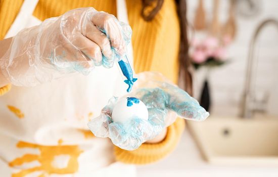 Beautiful brunette woman in yellow sweater and gray apron coloring easter eggs blue in the kitchen