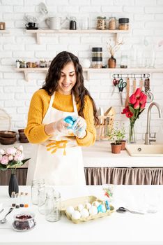 Beautiful brunette woman in yellow sweater and gray apron coloring easter eggs blue in the kitchen