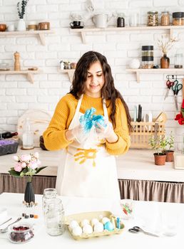 Beautiful brunette woman in yellow sweater and gray apron coloring easter eggs blue in the kitchen