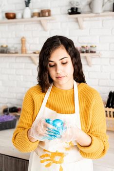 Beautiful brunette woman in yellow sweater and gray apron coloring easter eggs blue in the kitchen