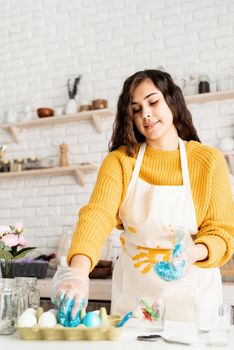 Beautiful brunette woman in yellow sweater and gray apron coloring easter eggs blue in the kitchen