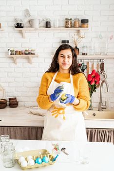 Beautiful brunette woman in yellow sweater and white apron coloring easter eggs in the kitchen