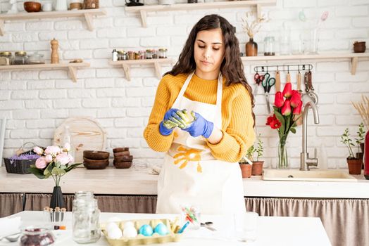 Beautiful brunette woman in yellow sweater and white apron coloring easter eggs in the kitchen