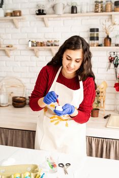 Beautiful brunette woman in red sweater and white apron coloring easter eggs in the kitchen