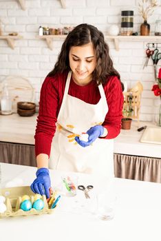 Beautiful brunette woman in red sweater and white apron coloring easter eggs in the kitchen