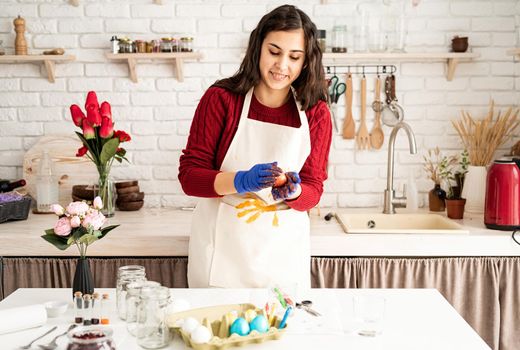 Beautiful brunette woman in red sweater and white apron coloring easter eggs in the kitchen
