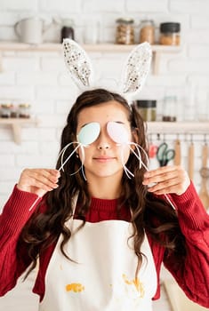 Beautiful funny brunette woman in red sweater and white apron wearing rabbit ears, covering eyes with easter eggs decorations