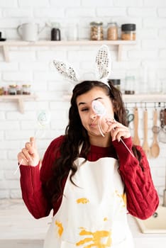 Beautiful funny brunette woman in red sweater and white apron wearing rabbit ears, covering eyes with easter eggs decorations