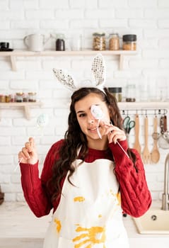 Beautiful funny brunette woman in red sweater and white apron wearing rabbit ears, covering eyes with easter eggs decorations