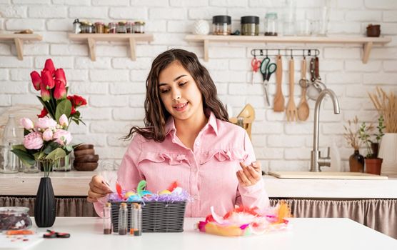 Beautiful smiling brunette woman in pink dress decorating colorful easter eggs with colorful feathers