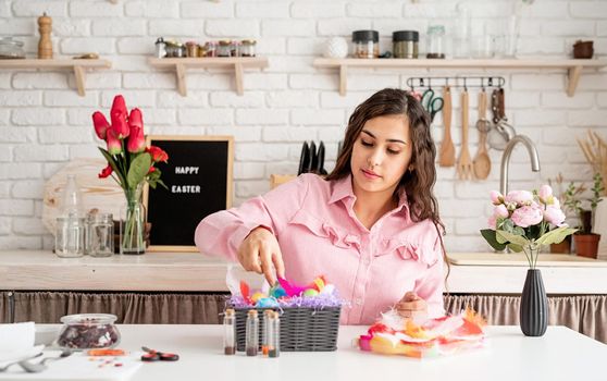 Beautiful smiling brunette woman in pink dress decorating colorful easter eggs with colorful feathers
