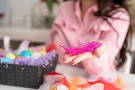 Beautiful smiling brunette woman in pink dress decorating colorful easter eggs with colorful feathers