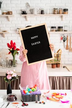 woman in pink dress holding black letter board with words Happy Easter, standing in the kitchen