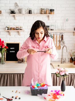 Beautiful brunette woman taking photo of decorated easter eggs using mobile phone in the kitchen