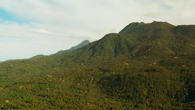 Mountains covered rainforest, trees and blue sky with clouds, aerial view. Camiguin, Philippines. Mountain landscape on tropical island with mountain peaks covered with forest. Slopes of mountains with evergreen vegetation.