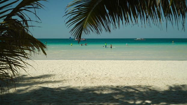 Tropical beach with tourists and clear blue sea through palm branches. Summer and travel vacation concept. Boracay, Philippines