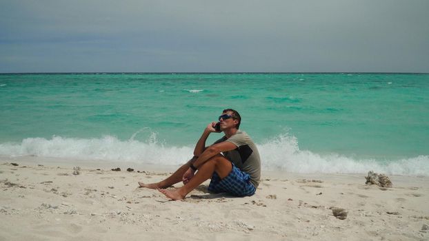 A man sits and talks on a cell phone on a tropical beach. Summer and travel vacation concept.