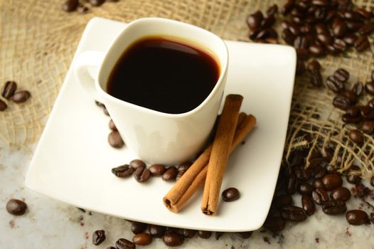A cup of espresso with a couple cinnamon sticks resting in a scene of roasted coffee beans.