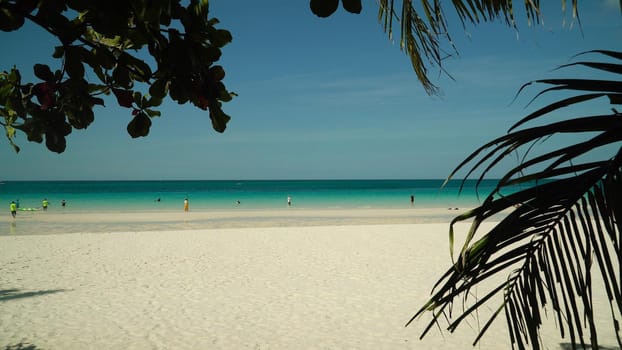 Tropical beach with tourists and clear blue sea through palm branches. Summer and travel vacation concept. Boracay, Philippines