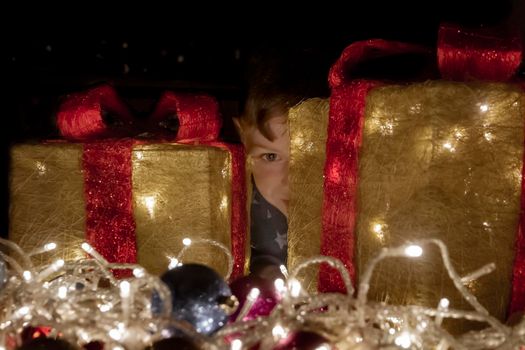 A cute blue-eyed, red-haired boy hiding behind some Christmas decorations with Christmas lights in the foreground