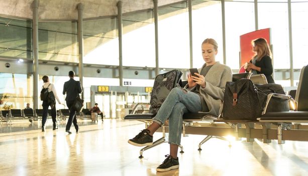 Casual blond young woman talking on cell phone while waiting to board a plane at airport departure gates.