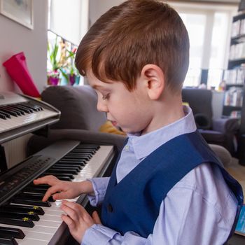 A cute, blue-eyed, red-haired, 4 years old boy playing the piano in a living room with a Christmas tree in thee background