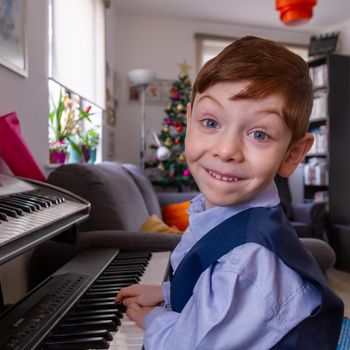 A cute, blue-eyed, red-haired, 4 years old boy playing the piano in a living room with a Christmas tree in thee background