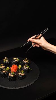 Hand with chopsticks wants to take custom sushi roll with black rice, crab meat, avocado, smoked salmon mousse, oar caviar, masago, shrimp cocktail, edible gold leaf, ginger, wasabi on black table