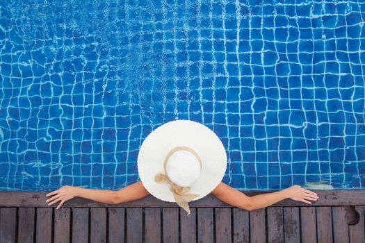 Beautiful woman sunbathing by the pool top view, summer vacation resort background. Girl sitting in swimming pool in big sunhat