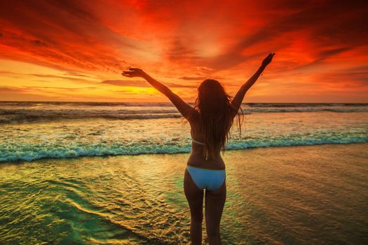 Woman in bikini walking with rised hands on the beach at sunset. Bali island, Indonesia