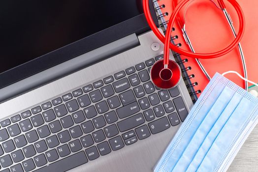 Virus pandemic and medical concept with a colorful red stethoscope coiled over a surgical face mask alongside a notebook on a wooden desk with computer keyboard viewed top down