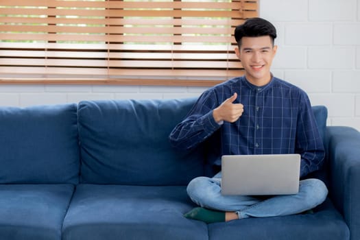 Young asian businessman smile and work from home with laptop computer online to internet on sofa in living room, freelance man using notebook on couch with comfort, new normal, lifestyle concept.