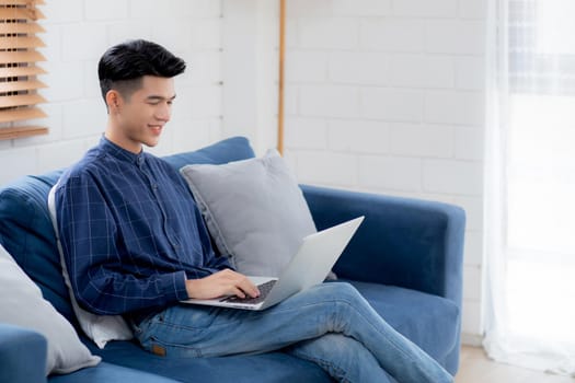 Young asian businessman smile and work from home with laptop computer online to internet on sofa in living room, freelance man using notebook on couch with comfort, new normal, lifestyle concept.