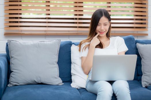 Beautiful young asian woman working on laptop computer and thinking idea on sofa at home, freelance girl sitting on couch using notebook to internet at living room, one person, lifestyle concept.