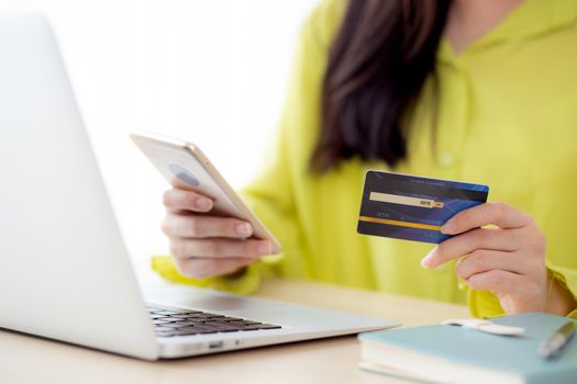 Closeup young asian business woman using smart phone and holding credit card while online shopping and payment with laptop computer on desk at home, female holding debit card, communication concept.