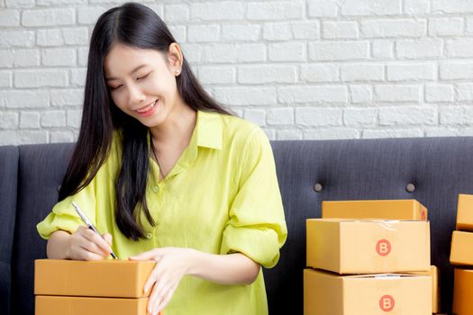 Young asian woman writing address on parcel box at home for delivery order to customer, shipping and logistic, merchant online and seller, business owner or SME, online shopping and e-commerce.