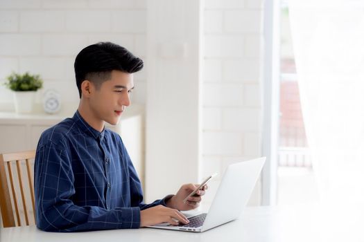Young asian man working laptop computer and reading smartphone on internet online on desk at home, freelance male smiling using phone with social media on table, business and communication concept.