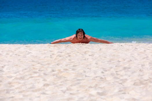 A young man with long black hair is lying on the sand by the sea, his arms spread wide. Blue waves, white sand.