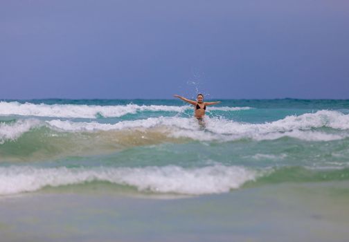 Young beautiful girl enjoys the sea and the waves, laughing raising her hands up in a black fashionable swimsuit.