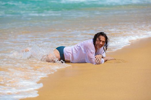 A young man with long black hair is lying on his stomach by the sea, he is getting wet from small waves.