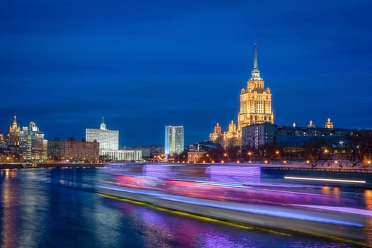 A ship with blurred lights moves along the river against the background of the evening city. Long exposure time.