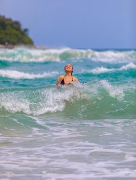 Young beautiful girl enjoys the sea and the waves, laughing raising her hands up in a black fashionable swimsuit.